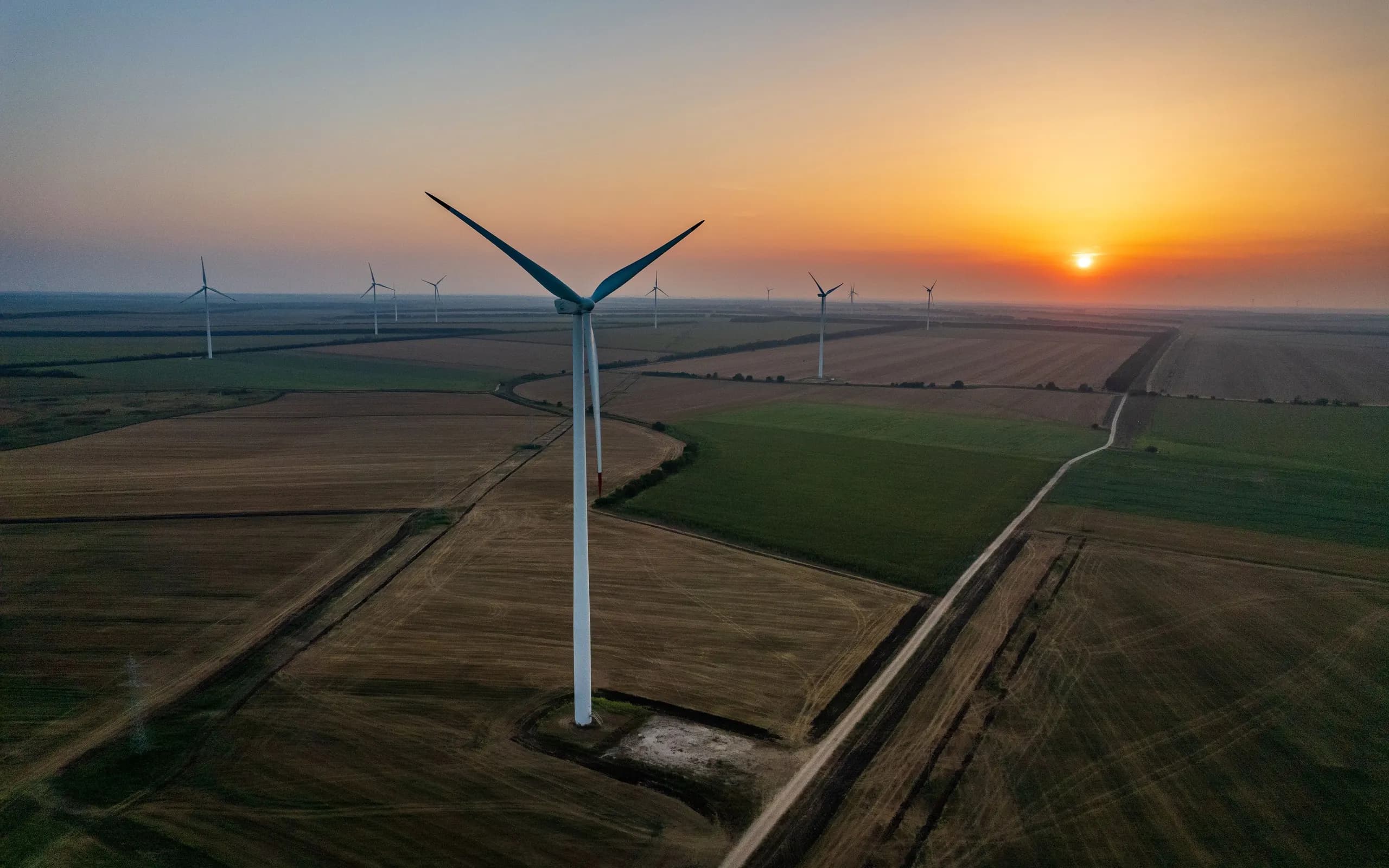 A wind farm at sunset, one turbine close and a dozen more across open fields